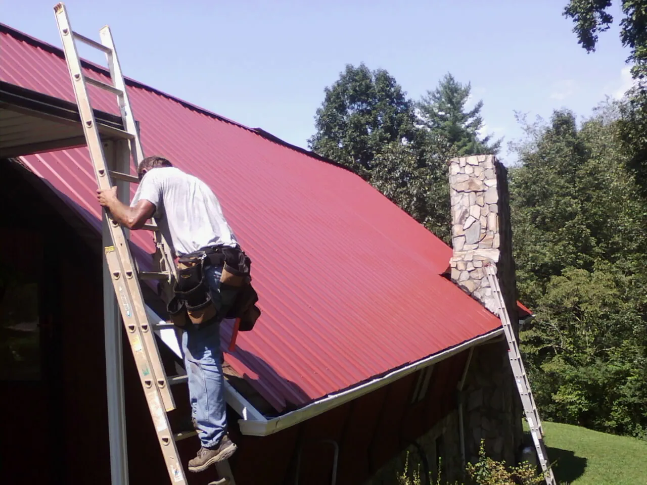 Roofer on a ladder beside a red metal roof and stone chimney — Soffit & Fascia Repair by Roofing Brothers in Maple Plain